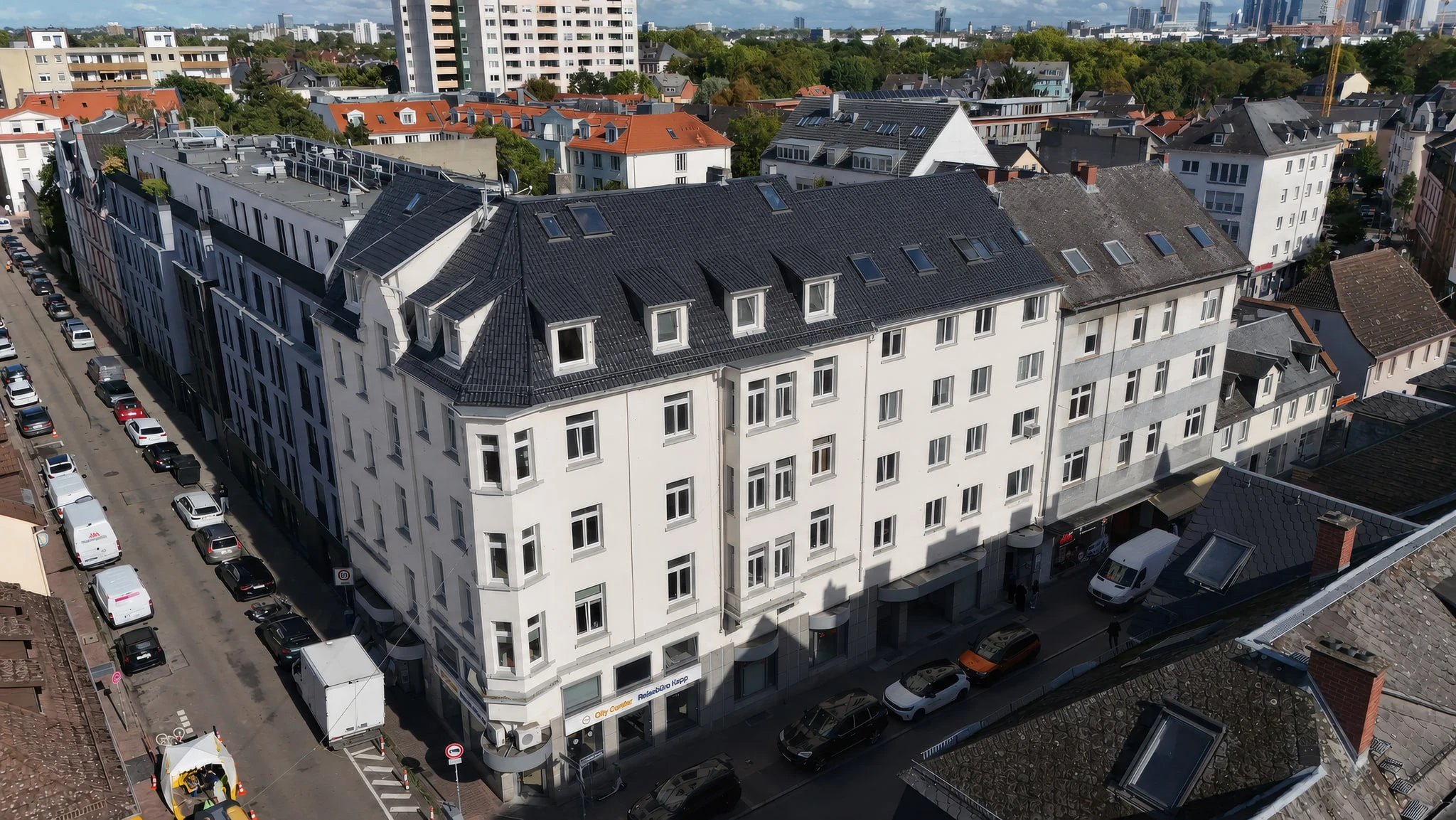 Das Ensemble CORNER 36 aus der Luft von schräg oben: die helle Straßenfassade mit der abgeschrägten Ecke und dem anschließenden Seitenflügel, darüber das dunkle Mansarddach mit Gauben und Dachflächenfenstern.