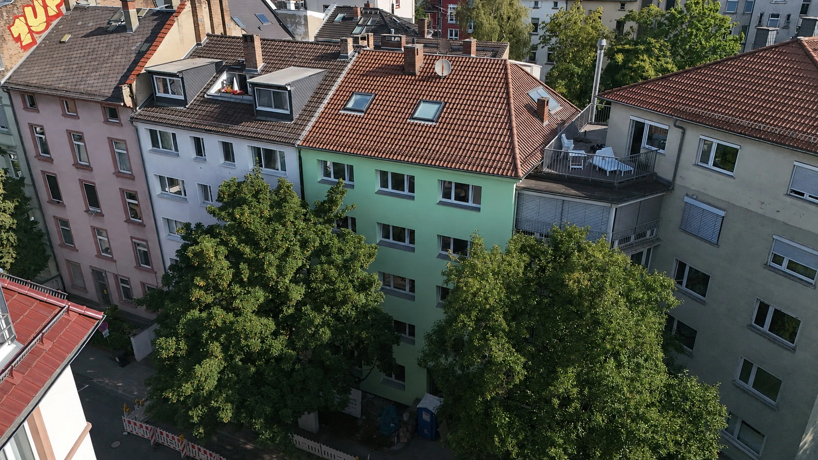 Das Projekt GREEN FOUR aus der Luft über der Straße: die mintgrüne Putzfassade mit vier Fensterachsen und rotem Ziegeldach, davor zwei ausladende Straßenbäume, links und rechts die angrenzenden Häuser.