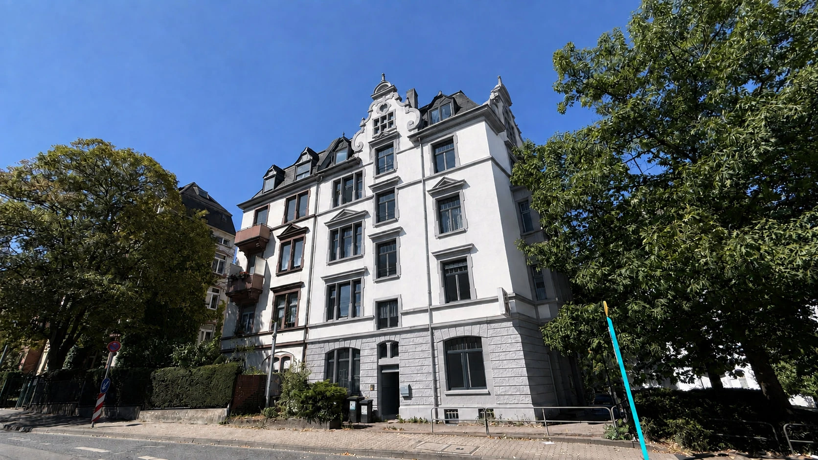 Das Projekt CROWN 78 von der Straße aus schräg unten: die weiße Fassade mit geschweiftem Giebel, Erker und dunklem Mansarddach vor blauem Himmel, links und rechts belaubte Bäume.