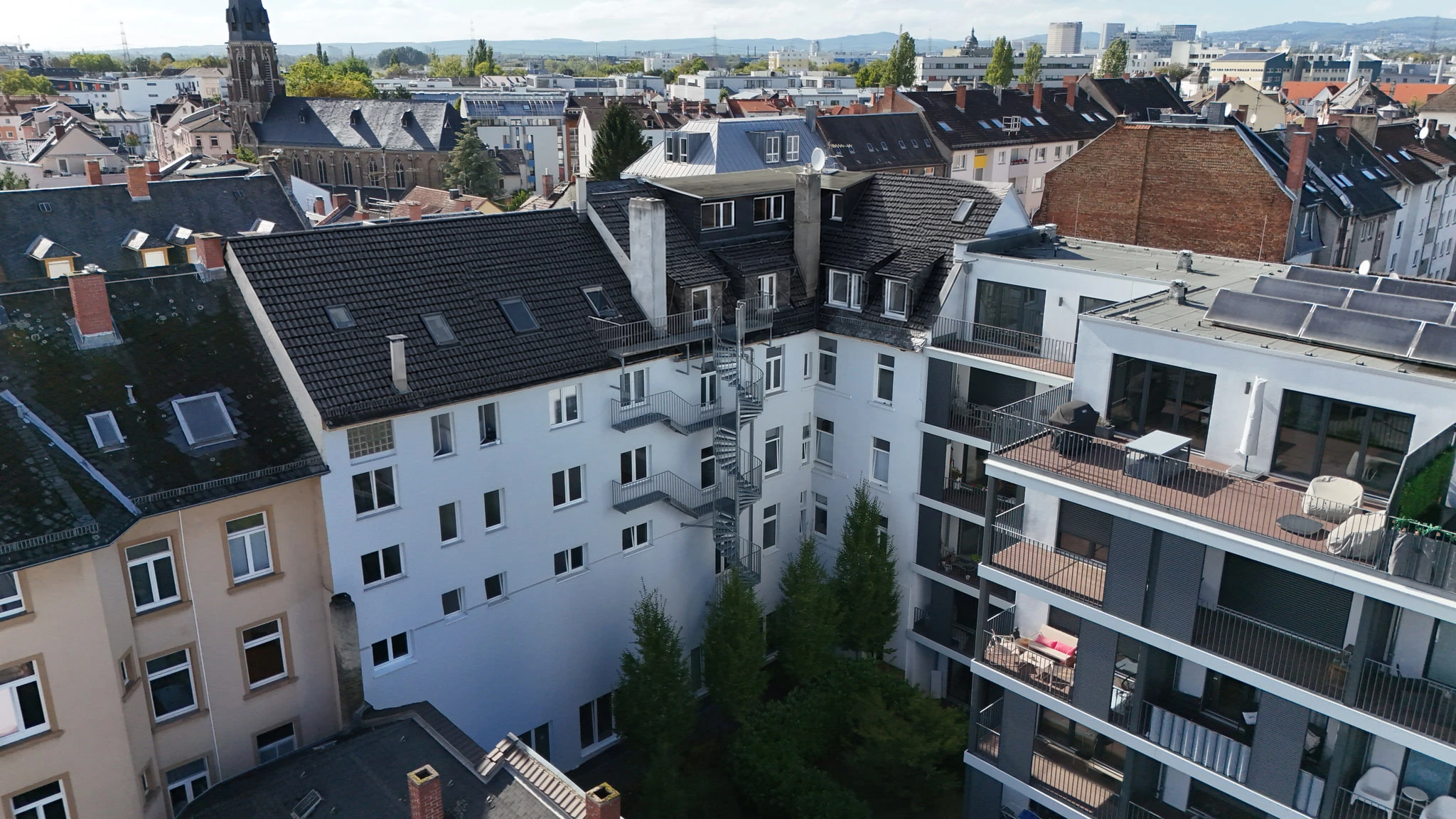 Die Hofseite von CORNER 36 aus der Luft: die weiße Rückfassade im rechten Winkel, davor eine stählerne Wendeltreppe mit Laubengängen und Bäume im Innenhof, im Hintergrund die Dächer des Viertels.