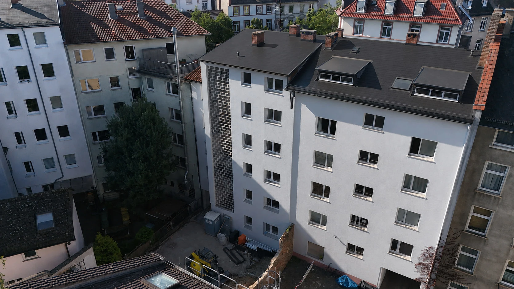 Die Hofseite des Projekts GREEN FOUR aus der Luft: hell verputzte Fassade mit dunkler Klinkerkante an der Gebäudeecke, dunkles Dach mit Gauben, im Hof davor Baugerät und Material.