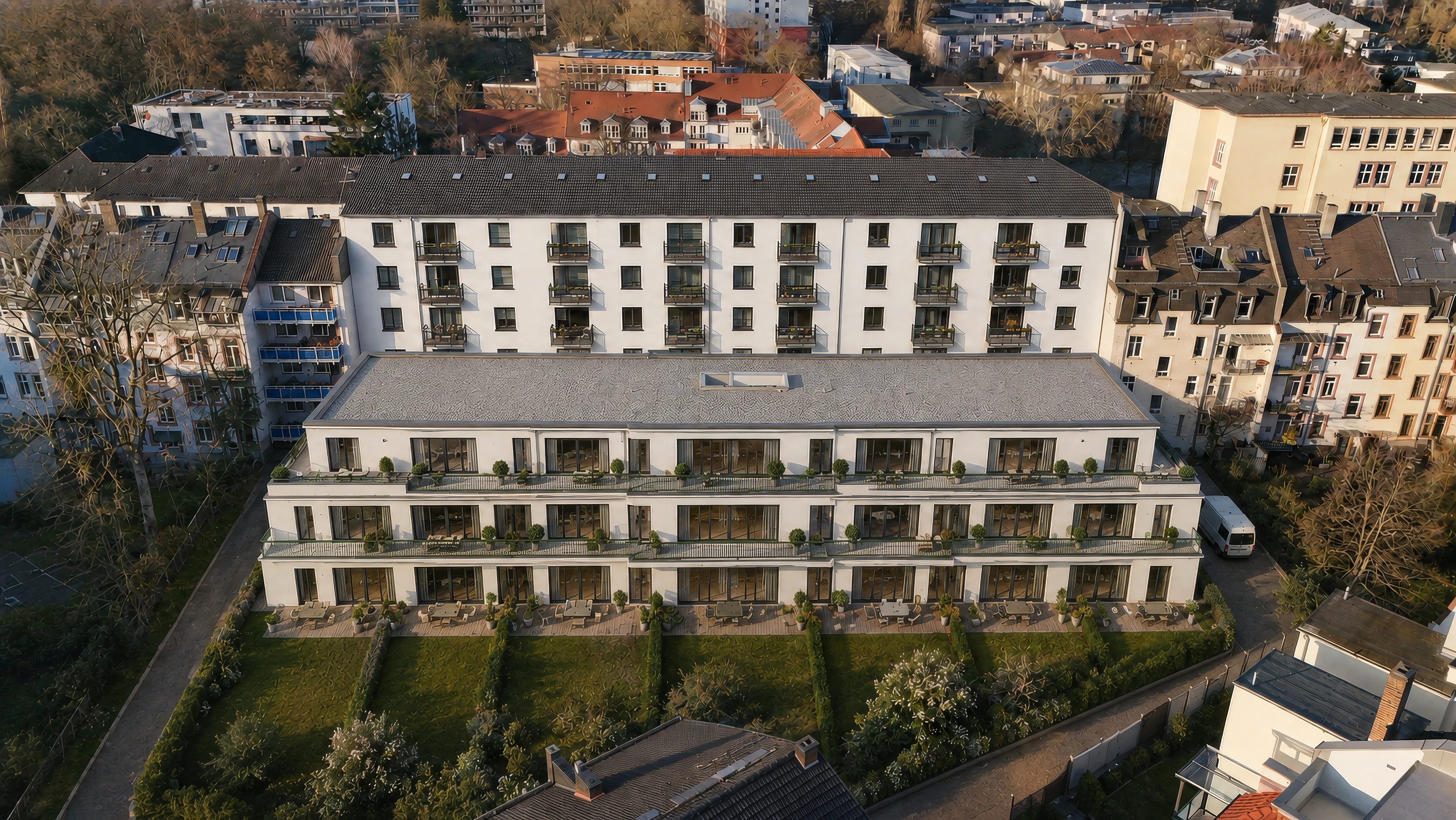 Der Neubau des Projekts ENSEMBLE 27 aus der Luft: ein dreigeschossiger heller Baukörper mit umlaufenden Terrassen und bepflanzten Gartenflächen im rückwärtigen Grundstücksbereich, dahinter die lange Bestandszeile mit Balkonen.