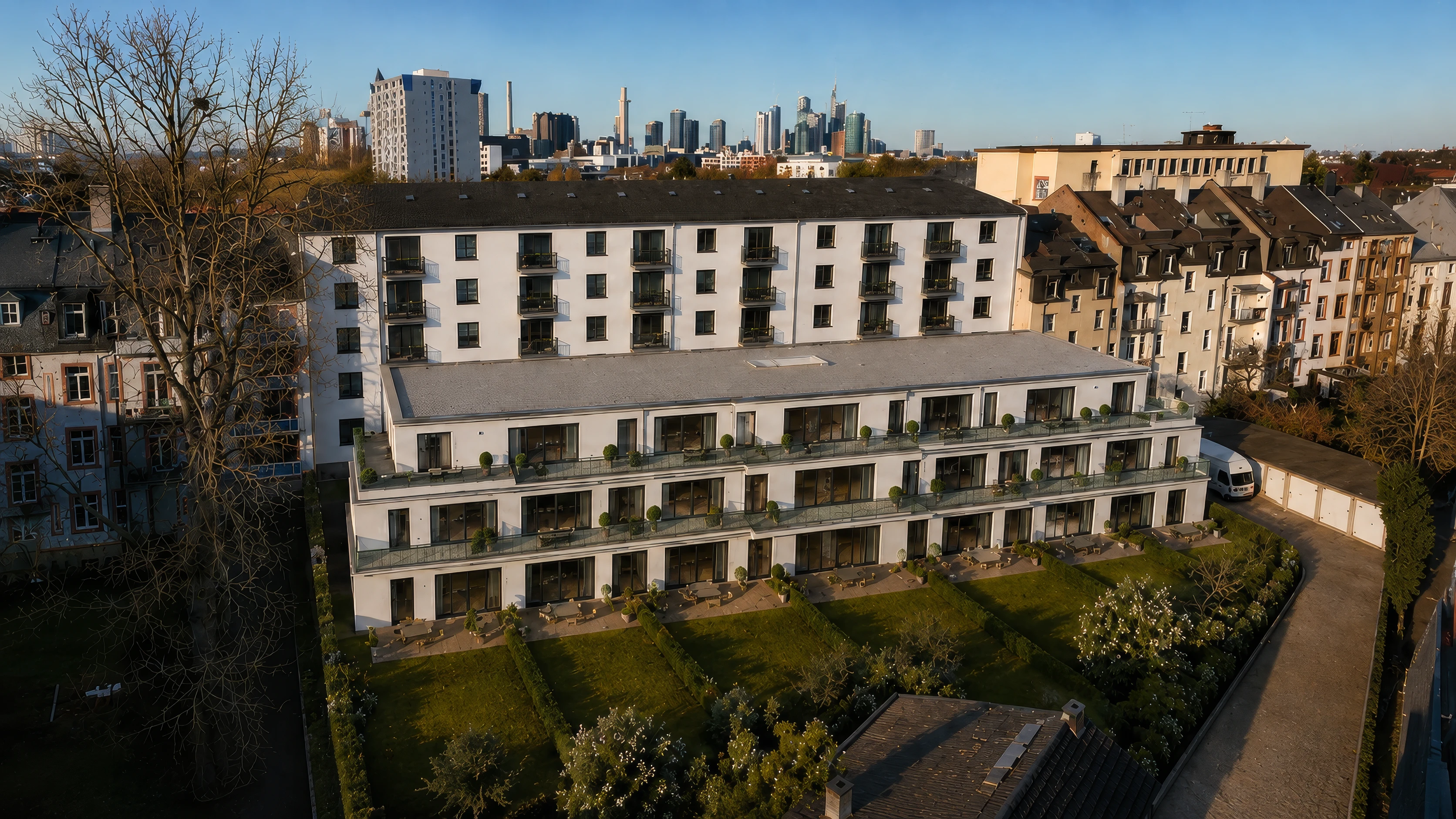 Dieselbe Anlage aus der anderen Richtung: der helle Neubau mit Terrassen und Rasenflächen vor der Bestandszeile, am Horizont die Hochhäuser der Frankfurter Skyline.