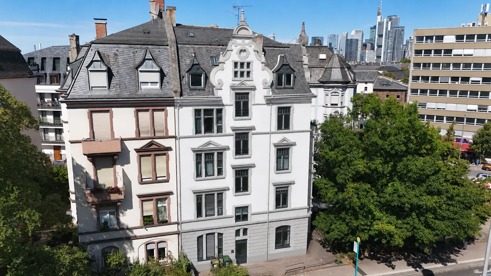Luftaufnahme des Projekts CROWN 78: ein weiß verputzter Gründerzeitbau mit geschweiftem Ziergiebel und dunklem Mansarddach an einer Straßenecke, davor hohe Laubbäume; im Hintergrund die Frankfurter Skyline.
