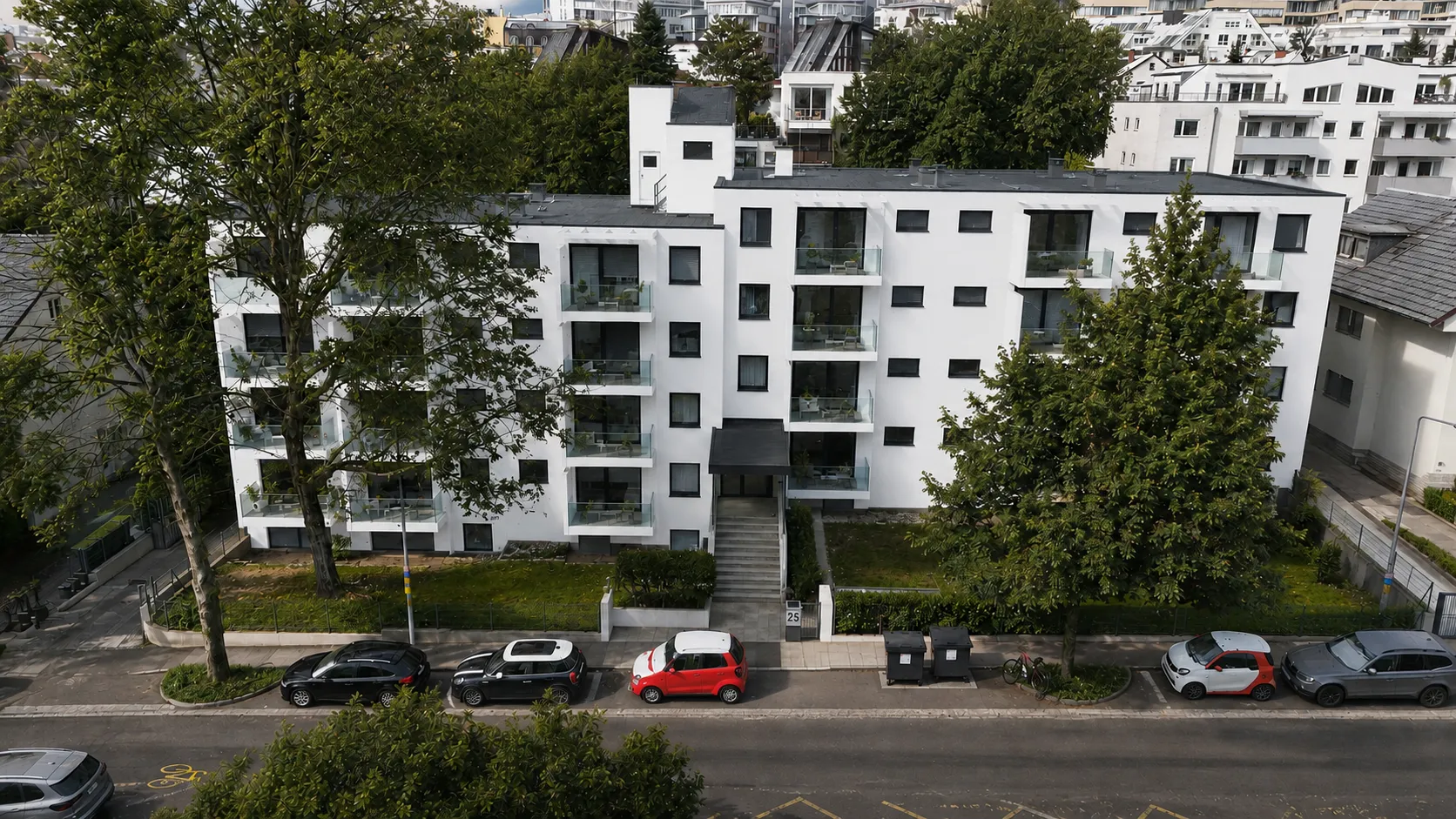 Luftaufnahme des Projekts SOUTH25: ein weißer Wohnbau mit dunklem Flachdach und umlaufenden Balkonen, davor hohe Laubbäume und die abschüssige Anliegerstraße mit parkenden Autos.