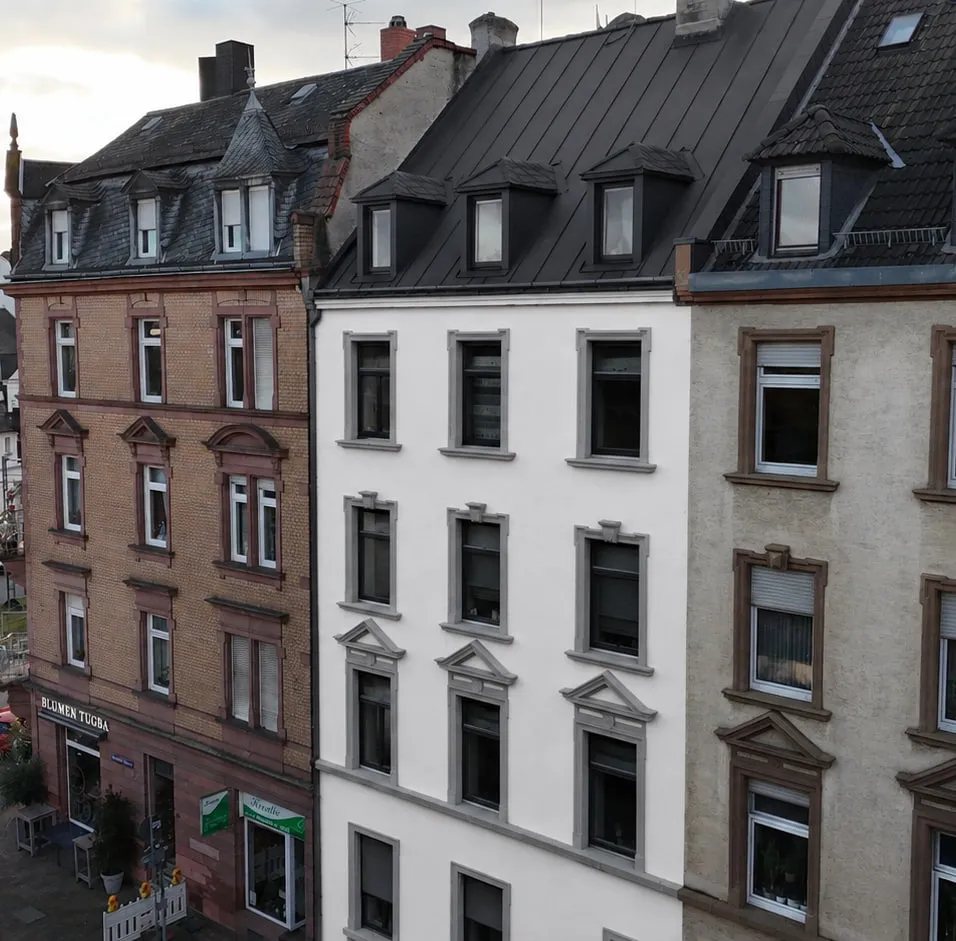 Das Projekt N°36 aus der Luft über der Straße: der weiße Altbau mit dunklem Dach in der Häuserzeile, davor die Fahrbahn mit Gehweg und Bäumen.