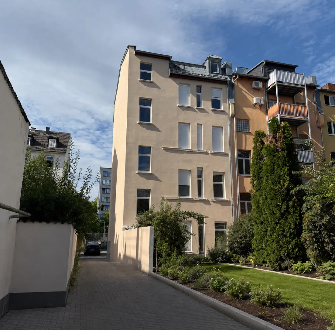 Die Gartenseite des Projekts WEST 27: sandfarbene Fassade mit hohen Fenstern über der begrünten Hoffläche, davor eine niedrige Mauer und dichte Sträucher.